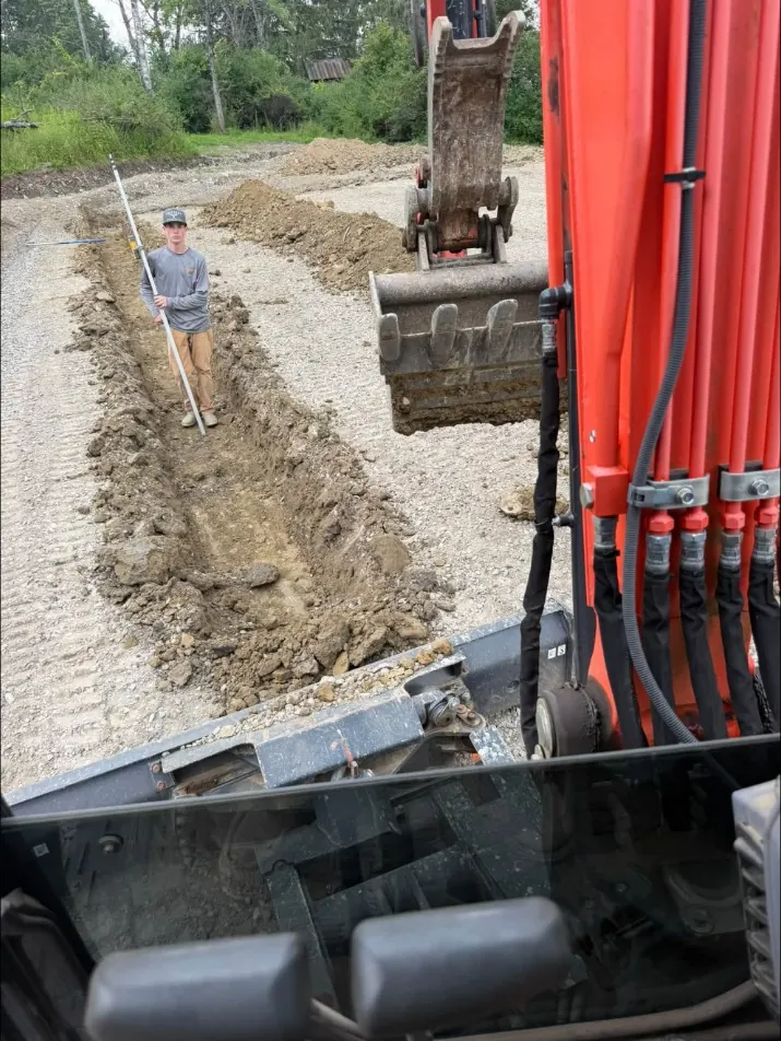 Digging a footing trench with the mini excavator while a crew member checks grade with a laser rod